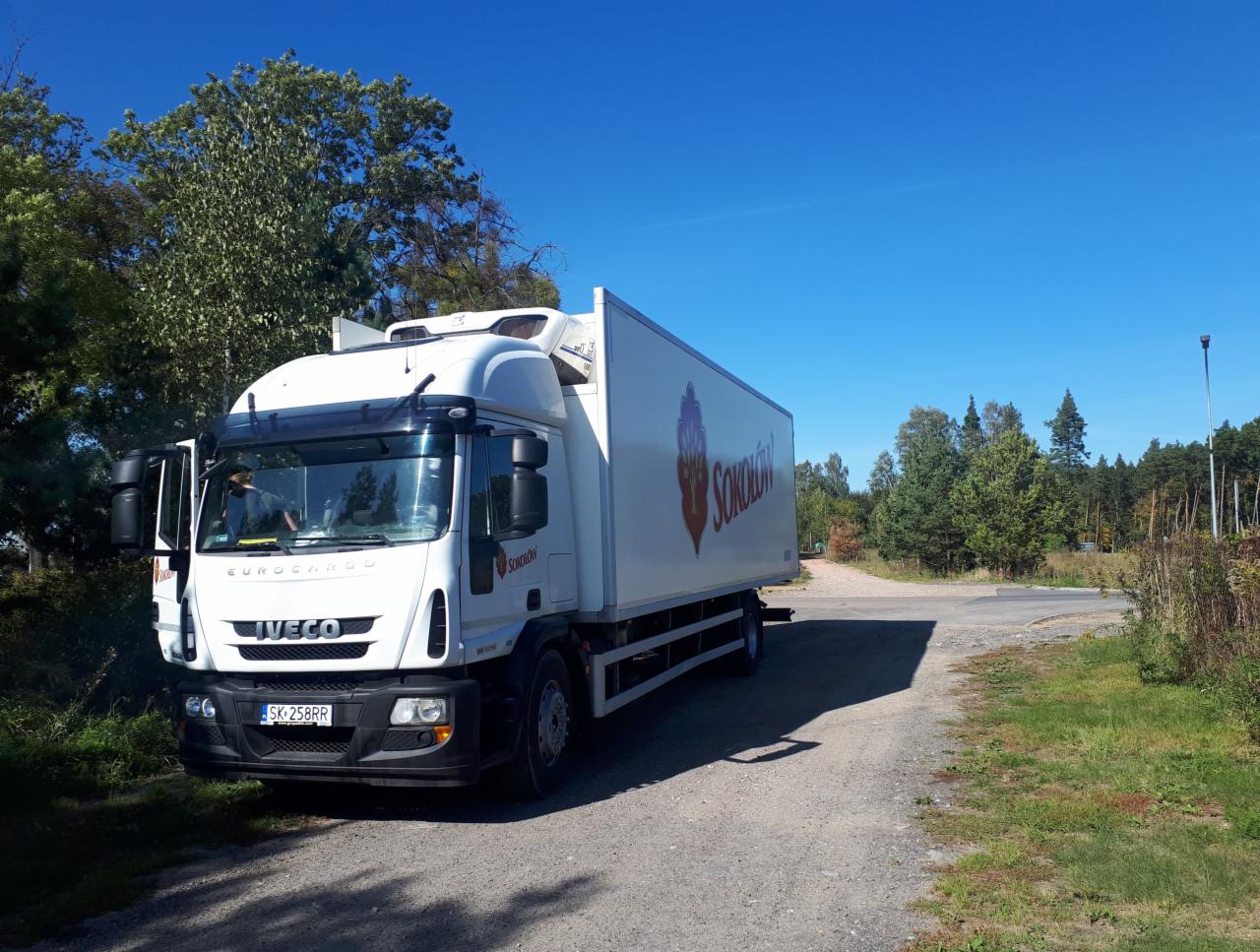 Biała ciężarówka chłodnia marki Iveco Eurocargo z otwartymi drzwiami kierowcy, zaparkowana na żwirowej drodze w słoneczny dzień, z logo firmy Sokolow na naczepie.