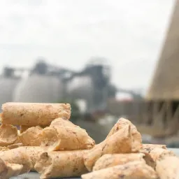 Stos granulatu opałowego z widoczną strukturą włókien, na tle rozmytym silosów i konstrukcji przemysłowych.