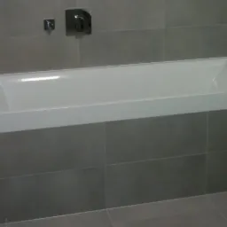 Wykończona łazienka z białą wanną obudowaną szarą glazurą i minimalistyczną armaturą.