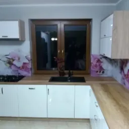 Nowoczesna kuchnia z białymi szafkami, drewnianym blatem i oknem w drewnianej ramie. Ściana nad blatem ozdobiona grafiką z motywem orchidei. Czarna kuchenka i zlew.