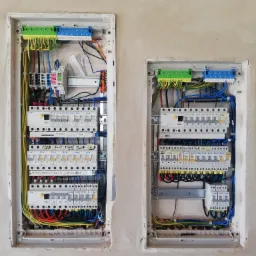 Dwie skrzynki elektryczne z zainstalowanymi bezpiecznikami i okablowaniem, zamontowane w surowej ścianie z widocznymi śladami po tynkowaniu.