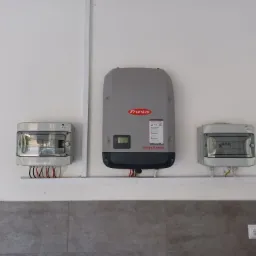 Szary falownik Fronius Symo zamontowany na białej ścianie, obok dwie skrzynki elektryczne z widocznymi przewodami, listwa montażowa i gniazdka elektryczne.