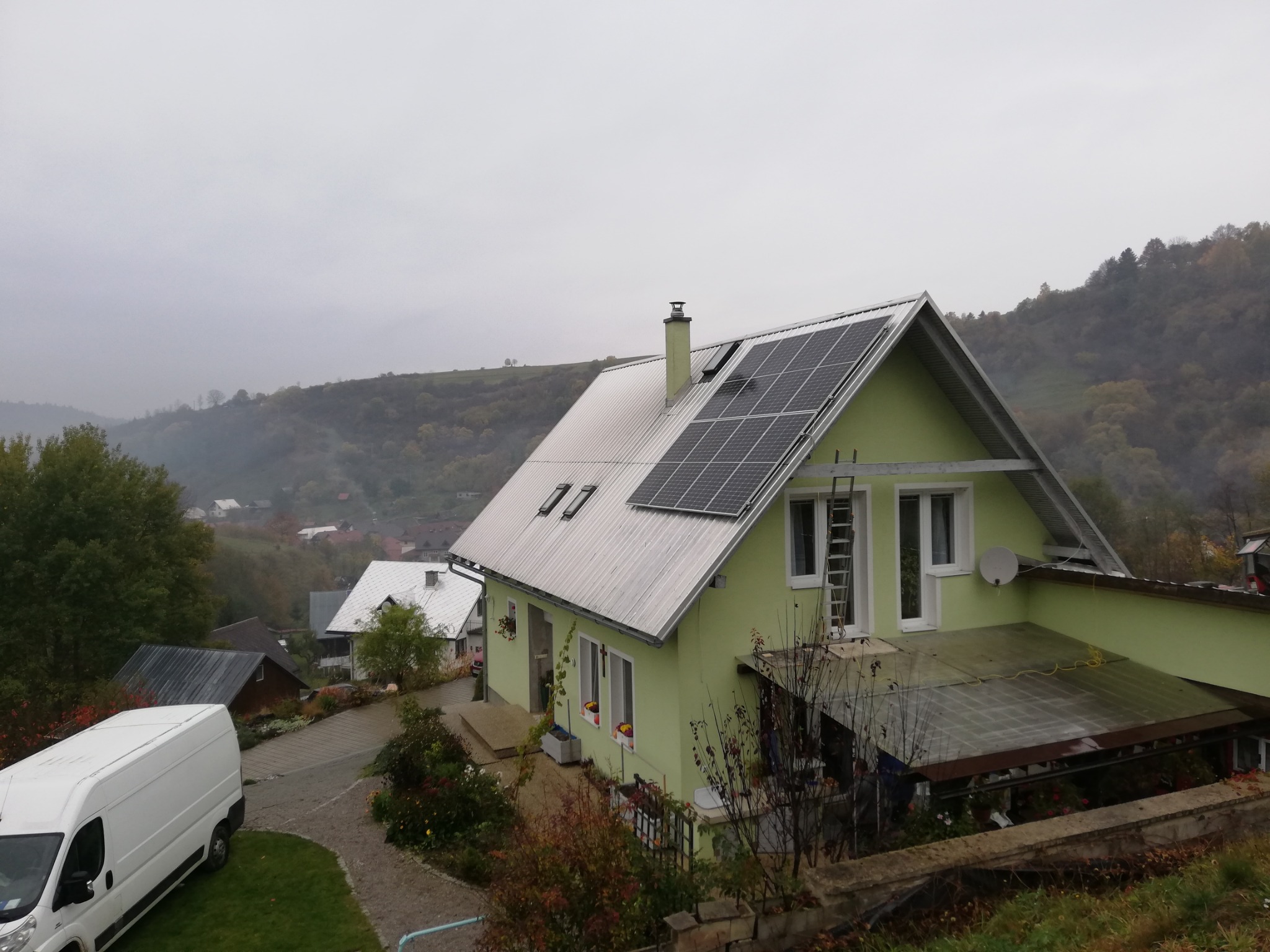 Zielony dom z panelami fotowoltaicznymi na dachu, widok z góry, w tle zalesione wzgórza spowite mgłą.
