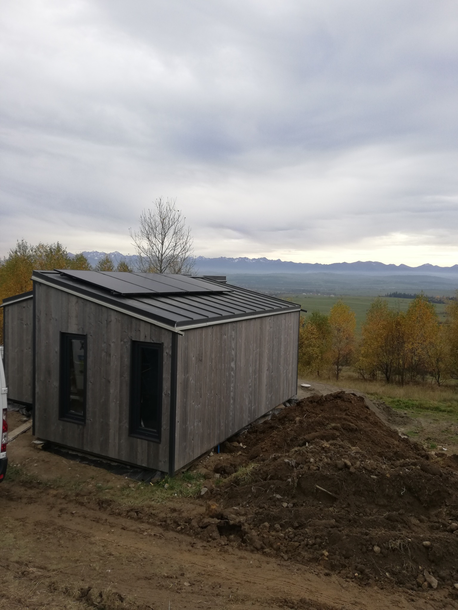 Nowoczesny, drewniany dom modułowy z panelami słonecznymi na dachu, w tle rozległy krajobraz z górami i jesiennymi drzewami. Widoczny stos ziemi przy budynku.