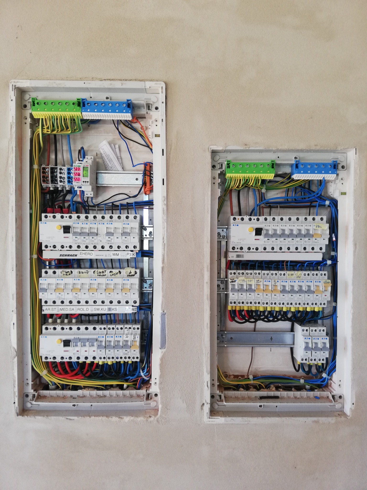 Dwie skrzynki elektryczne z zainstalowanymi bezpiecznikami i okablowaniem, zamontowane w surowej ścianie z widocznymi śladami po tynkowaniu.