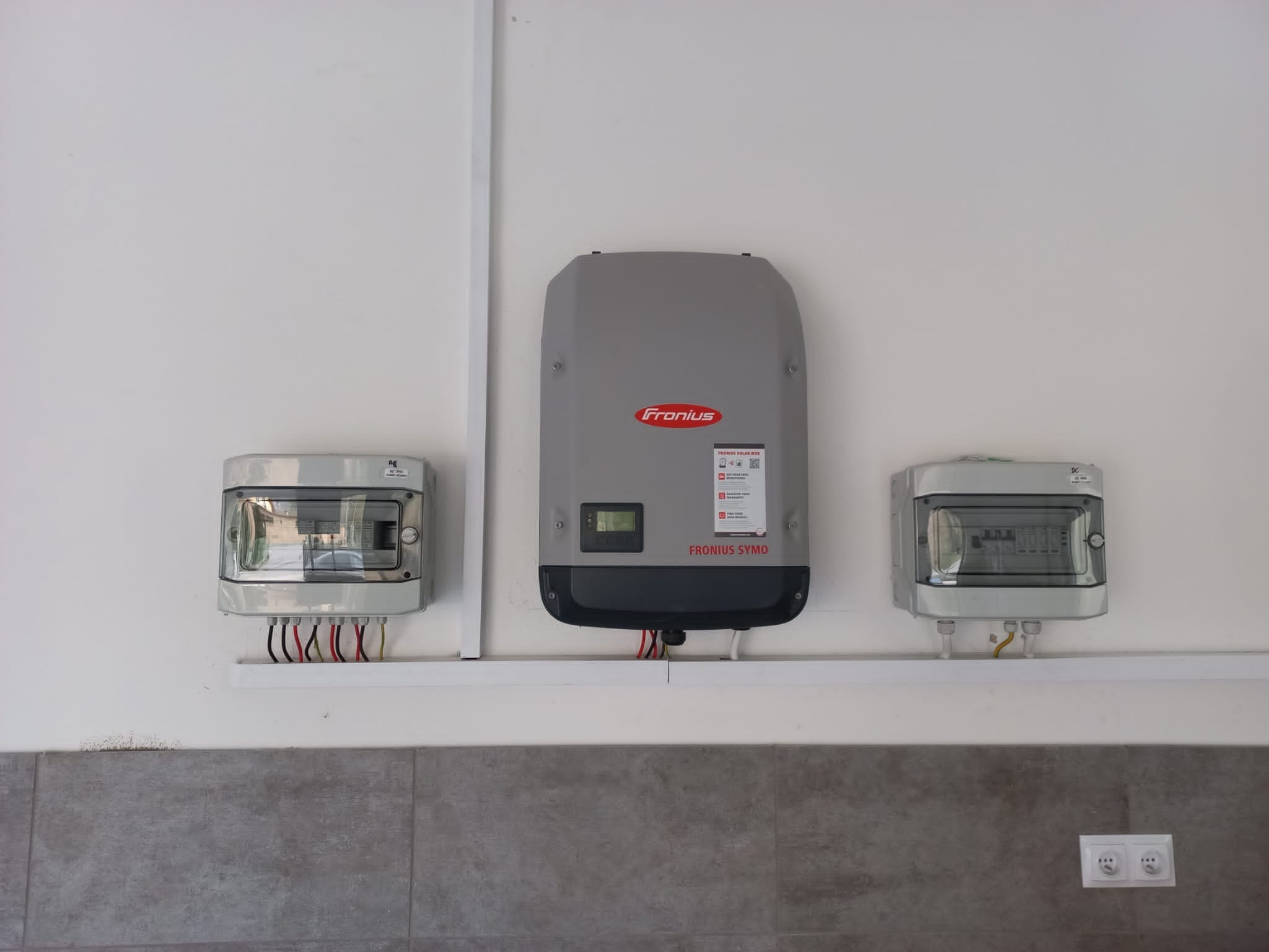 Szary falownik Fronius Symo zamontowany na białej ścianie, obok dwie skrzynki elektryczne z widocznymi przewodami, listwa montażowa i gniazdka elektryczne.