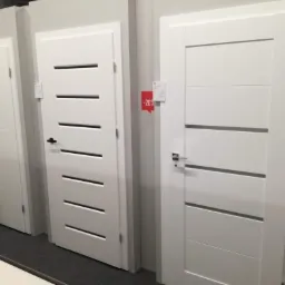 Wystawa trzech białych drzwi z poziomymi, prostokątnymi przeszkleniami, widoczne r&oacute;żne modele klamek i naklejka promocyjna -20%.