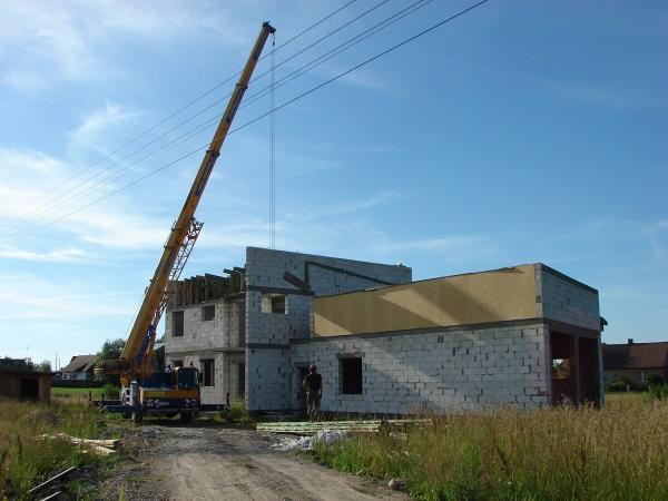 Budowa domu murowanego z użyciem dźwigu, widoczne bloczki betonowe i drewniana konstrukcja dachu w trakcie montażu.