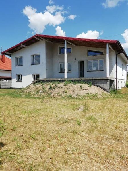 Dwukondygnacyjny dom z czerwonym dachem w trakcie budowy, z widocznym nasypem ziemi przed budynkiem i suchą trawą na pierwszym planie.