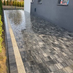 KONSBRUK - Świeżo ułożona, mokra nawierzchnia z ciemnoszarej kostki brukowej z jasnym, ozdobnym pasem, przylegająca do ściany budynku w odcieniach szarości i bieli.