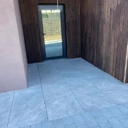 Nowoczesny taras wyłożony szarymi płytami ceramicznymi, przylegający do ściany budynku w kolorze pudrowego różu i drewnianej elewacji z ciemnego drewna. W tle widoczne drzwi wejściowe...