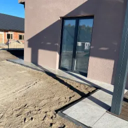 Przygotowanie terenu pod taras z płyt betonowych przy nowym domu, widoczne ślady opon na ziemi