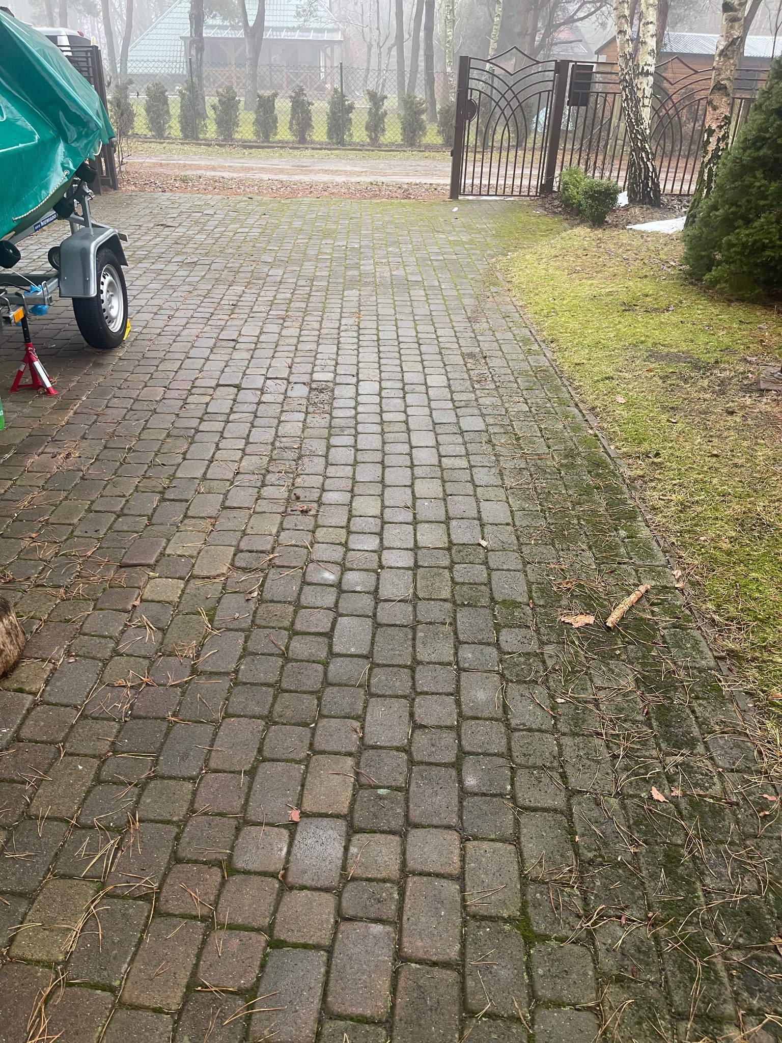 Ułożona kostka brukowa na podjeździe w pochmurny dzień, widoczne igliwie i przyczepa z łodzią w tle.