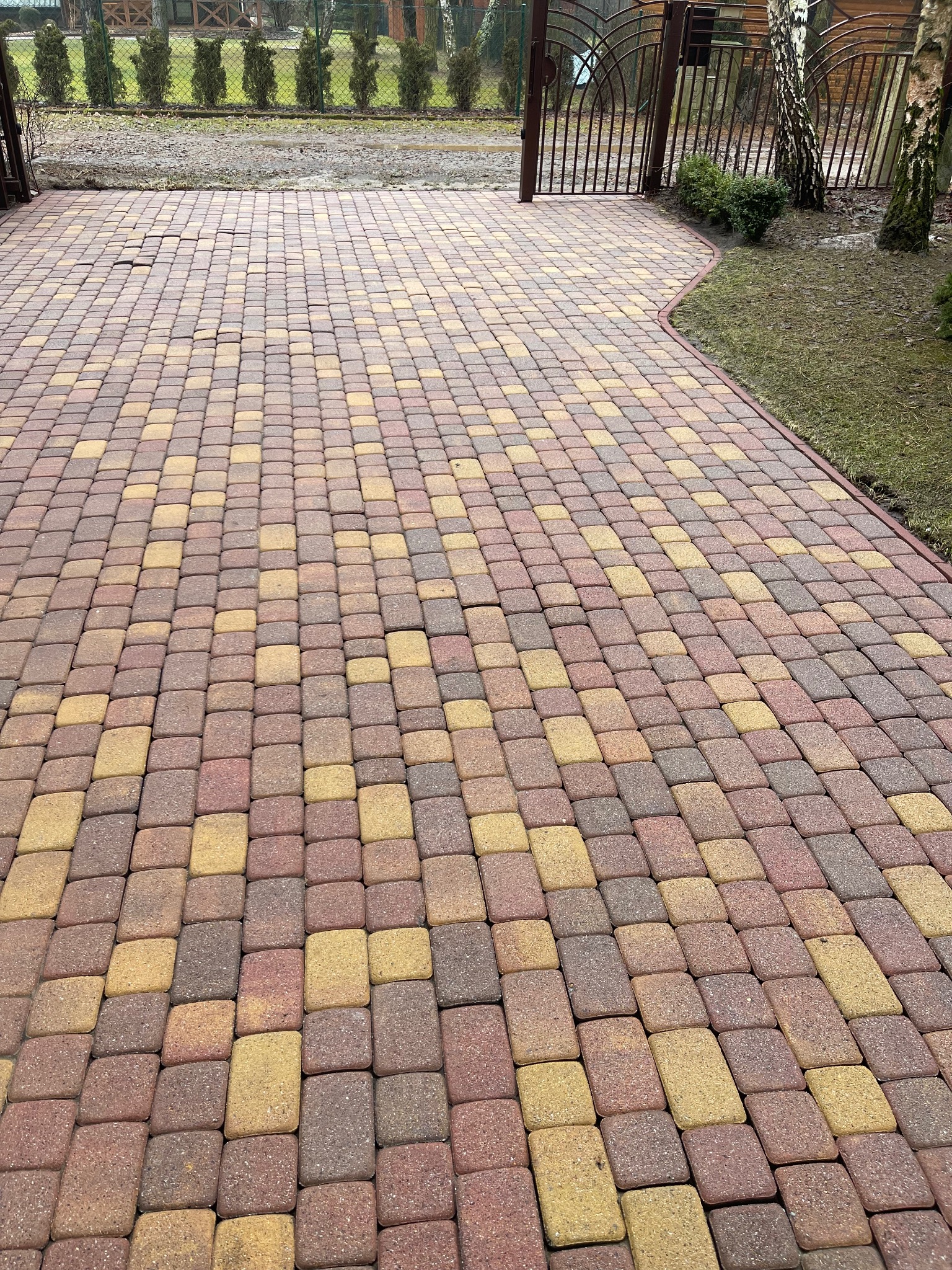 Ułożona, kolorowa kostka brukowa prowadząca do bramy wjazdowej, z widocznym obrzeżem i trawnikiem po prawej stronie.