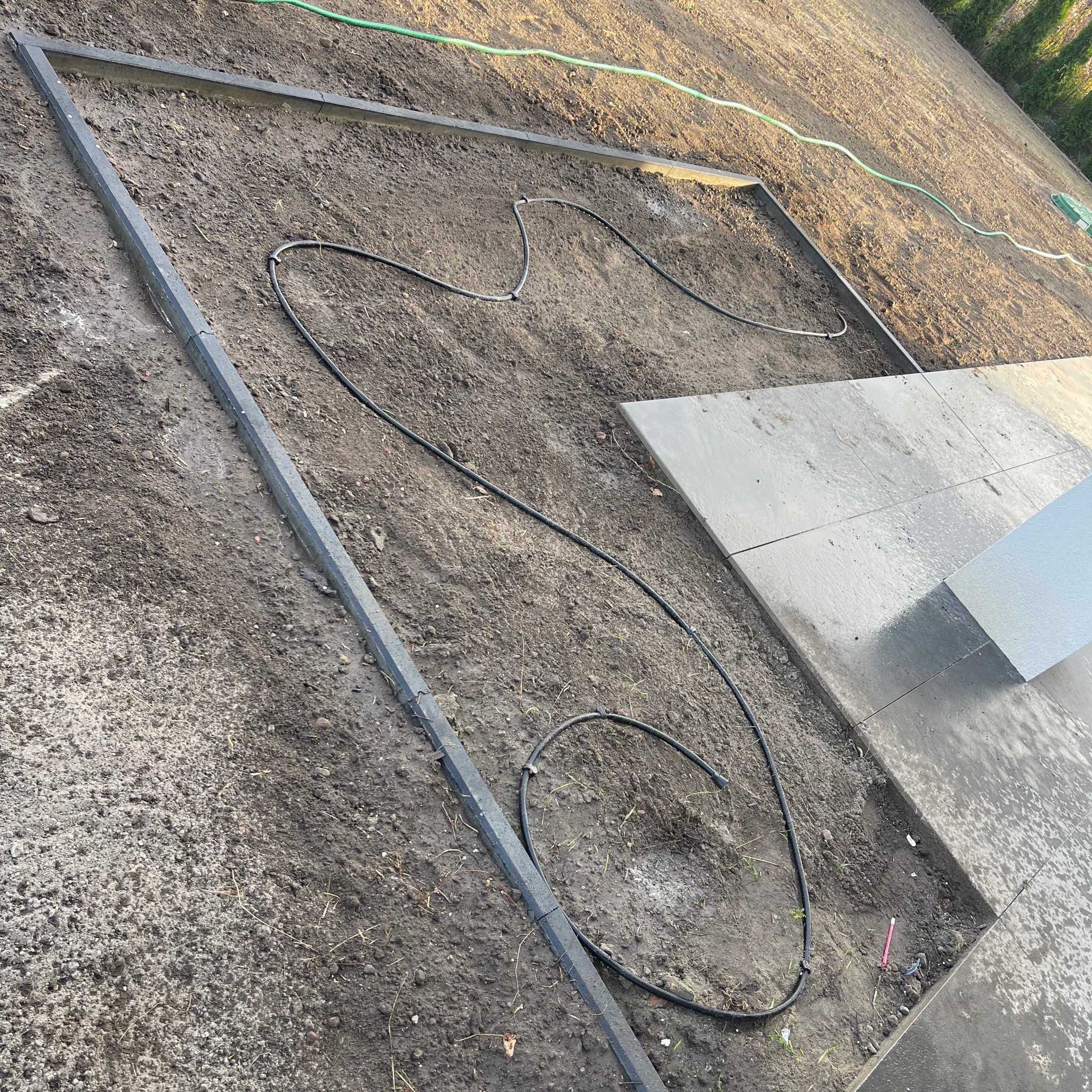 Przygotowany teren pod montaż nawadniania i ułożenie płyt, widoczne obrzeża, ziemia, węże nawadniające i częściowo ułożone płyty.