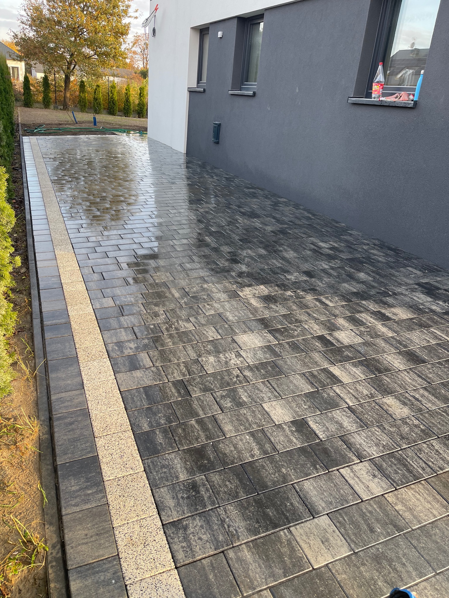 Świeżo ułożona, mokra nawierzchnia z ciemnoszarej kostki brukowej z jasnym, ozdobnym pasem, przylegająca do ściany budynku w odcieniach szarości i bieli.