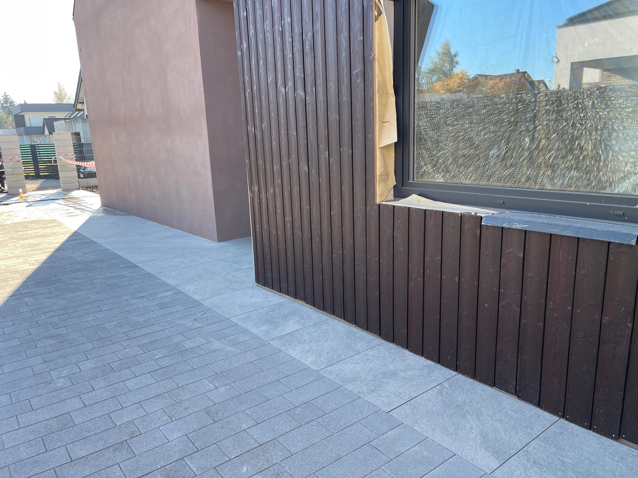 Nowoczesna posesja z szarej kostki brukowej prowadzącej do budynku z drewnianą elewacją i dużym oknem, zabezpieczonym folią malarską.