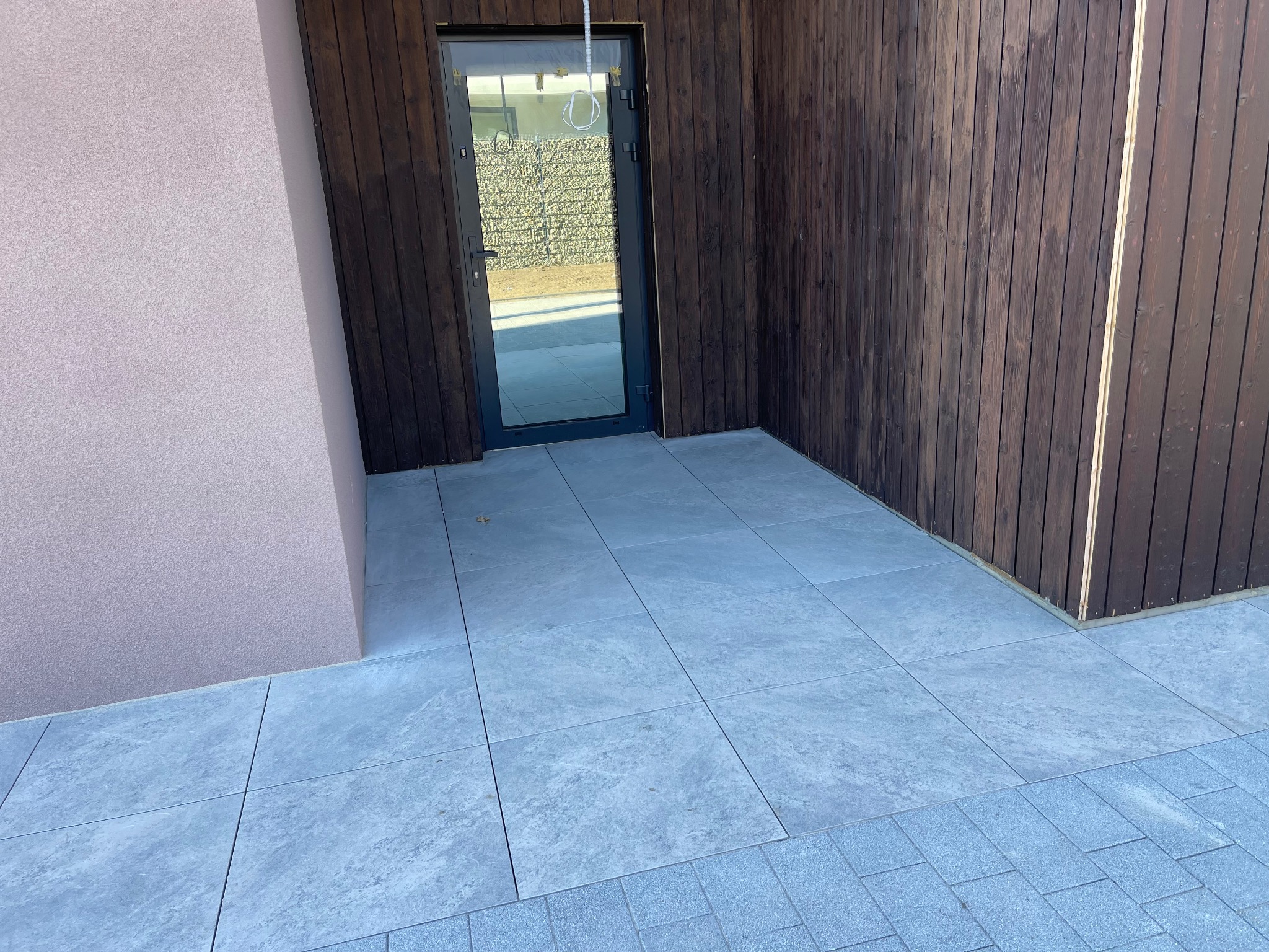 Nowoczesny taras wyłożony szarymi płytami ceramicznymi, przylegający do ściany budynku w kolorze pudrowego różu i drewnianej elewacji z ciemnego drewna. W tle widoczne drzwi wejściowe...