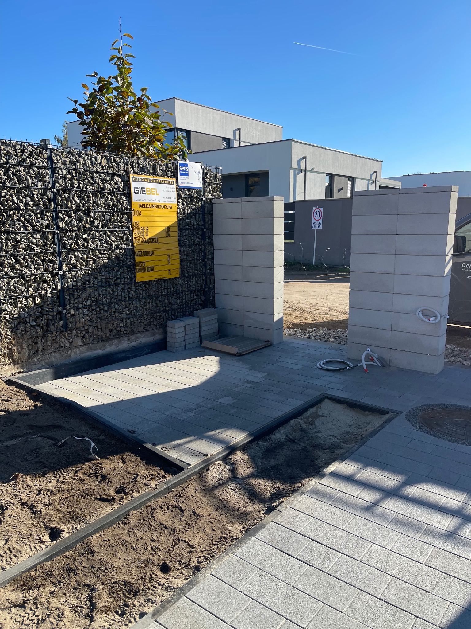 Układanie szarej kostki brukowej na posesji z gabionami, widoczne przygotowane podłoże, obrzeża, słupki wejściowe z pustaków, tablica informacyjna budowy i znak ograniczenia prędkości. Częściowo...