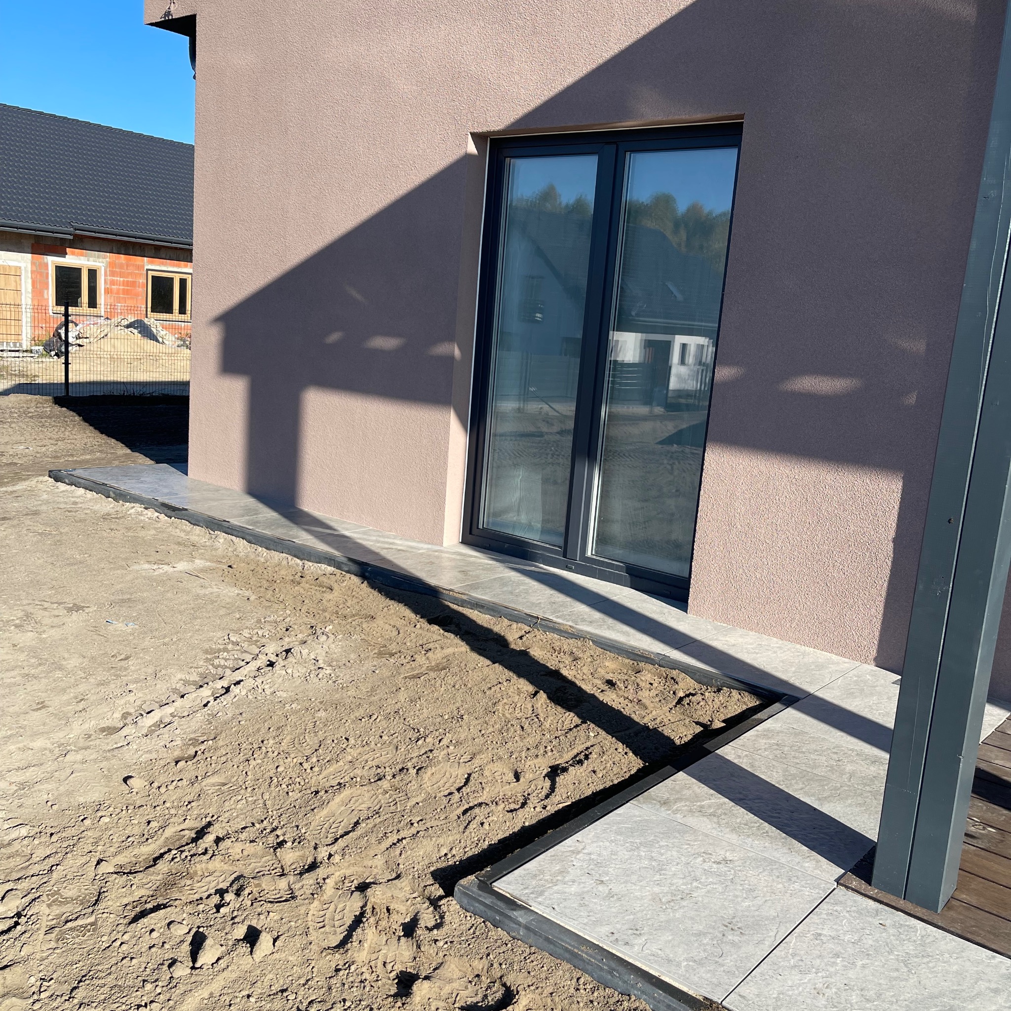 Przygotowanie terenu pod taras z płyt betonowych przy nowym domu, widoczne ślady opon na ziemi