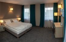 Pokój hotelowy z dwoma łóżkami z białymi pościelami, biurkiem, krzesłem i zasłonami w odcieniach bieli i turkusu, oświetlony lampkami nocnymi. Na biurku szklanka z wodą.