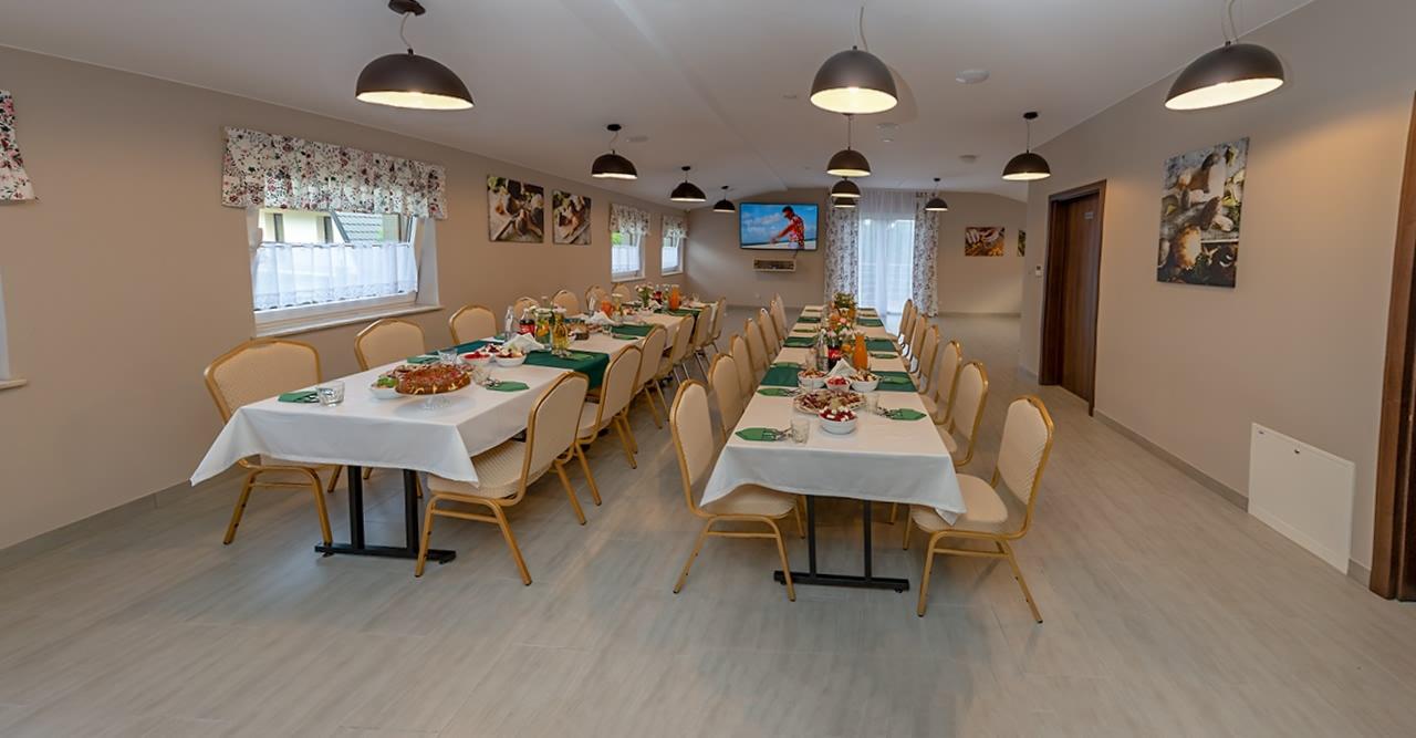 Sala bankietowa z długimi stołami nakrytymi białymi obrusami, przygotowana na przyjęcie, z dekoracjami, napojami i przekąskami, oświetlona wiszącymi lampami w kształcie kopuł.