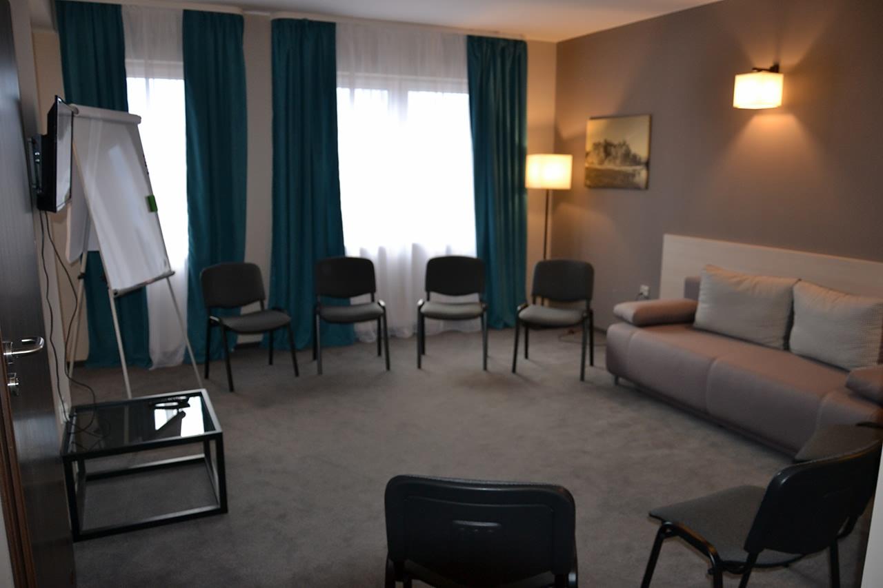 Pokój terapeutyczny z krzesłami ustawionymi w kręgu, tablicą suchościeralną, szarą sofą i stonowaną kolorystyką ścian.