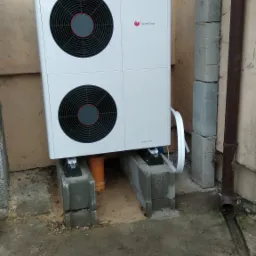 Biała pompa ciepła Saunier Duval GeniaAir Split, zamontowana na betonowych blokach przy ścianie budynku z widocznym oknem i rurą spustową, na ziemi ślady błota.