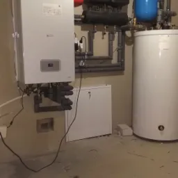 Instalacja grzewcza w piwnicy z białym kotłem, zasobnikiem ciepłej wody, czerwonym naczyniem przeponowym i czarnymi rozdzielaczami oraz szarymi rurami.