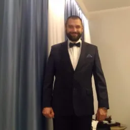 Uśmiechnięty mężczyzna w ciemnym garniturze i muszce, stojący na tle zasłon i włączonego telewizora w pokoju.