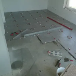 Układanie szarych płytek podłogowych w pomieszczeniu, widoczne poziomice, krzyżyki dystansowe, przyssawki do płytek i narzędzia glazurnicze, fragment nieukończonej podłogi.