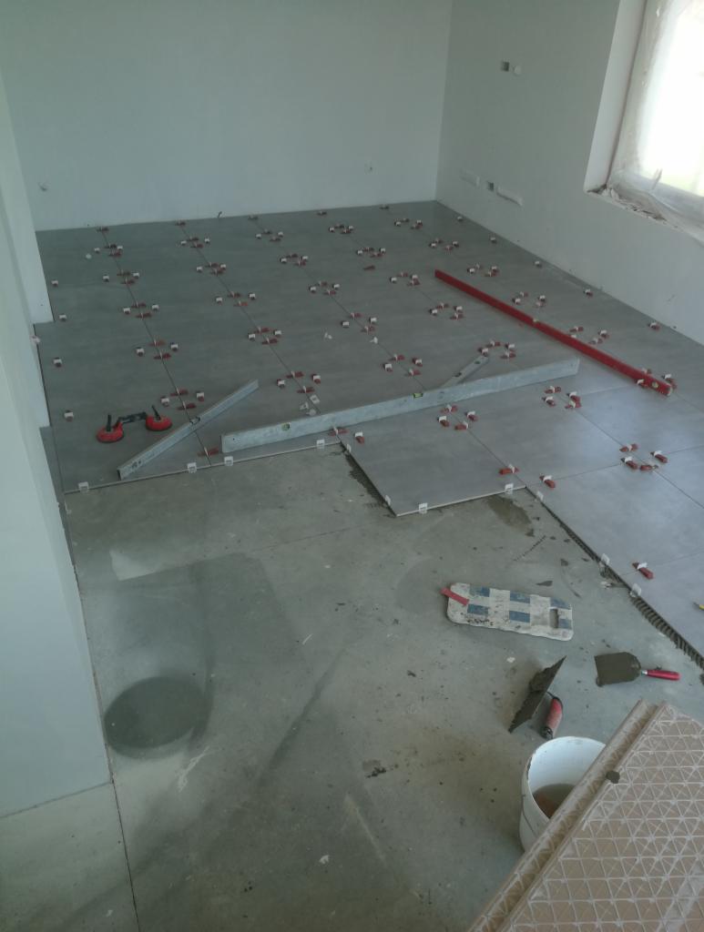 Układanie szarych płytek podłogowych w pomieszczeniu, widoczne poziomice, krzyżyki dystansowe, przyssawki do płytek i narzędzia glazurnicze, fragment nieukończonej podłogi.