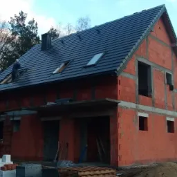 Budowa domu z czerwonej cegły: murarz pracuje na dachu z czarną dachówką, montując okna dachowe, widoczne rusztowania i materiały budowlane na placu budowy