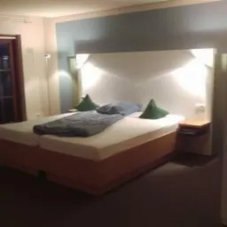 Pokój hotelowy z podwójnym łóżkiem, zielonymi poduszkami i przesuwnymi drzwiami balkonowymi, oświetlony ciepłym światłem lamp nocnych.