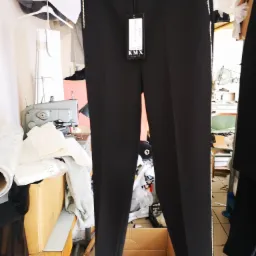 Czarne spodnie damskie z lampasami z cyrkonii wiszące na wieszaku w pracowni krawieckiej, w tle maszyna do szycia i materiały.