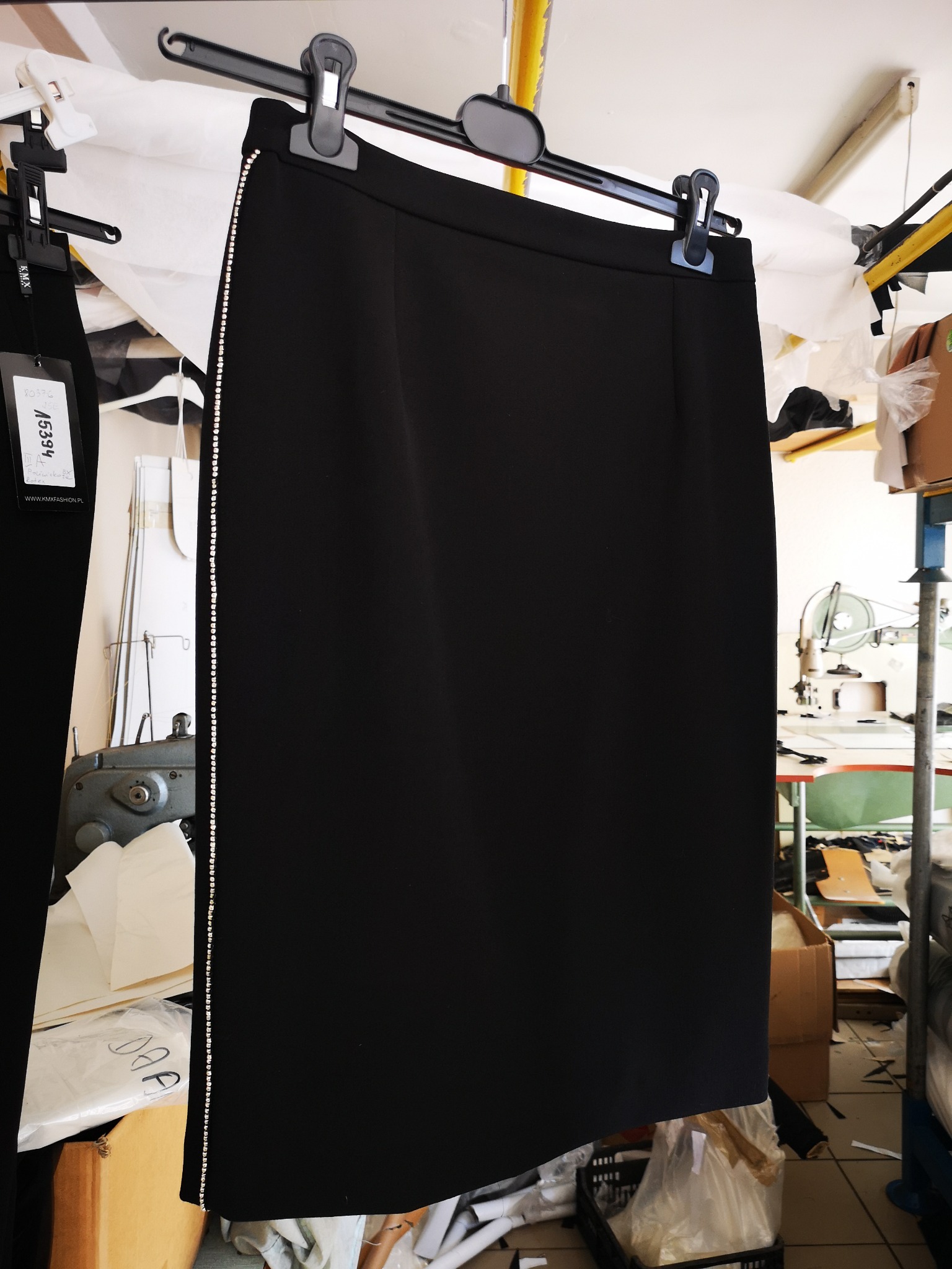 Czarna spódnica ołówkowa z ozdobnym paskiem kryształków na boku, wisząca na wieszaku w szwalni, widoczny fragment maszyny do szycia w tle.
