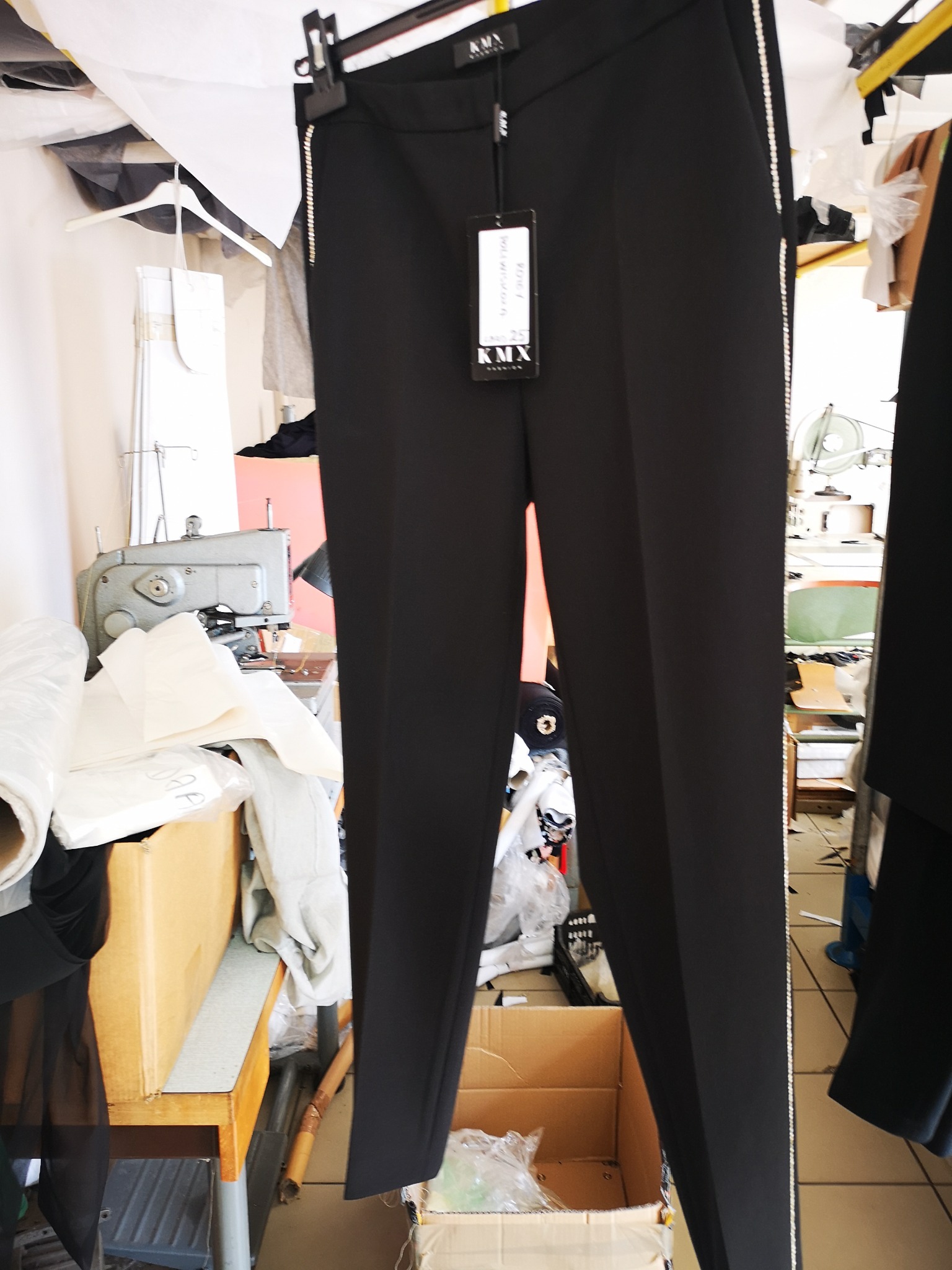 Czarne spodnie damskie z lampasami z cyrkonii wiszące na wieszaku w pracowni krawieckiej, w tle maszyna do szycia i materiały.