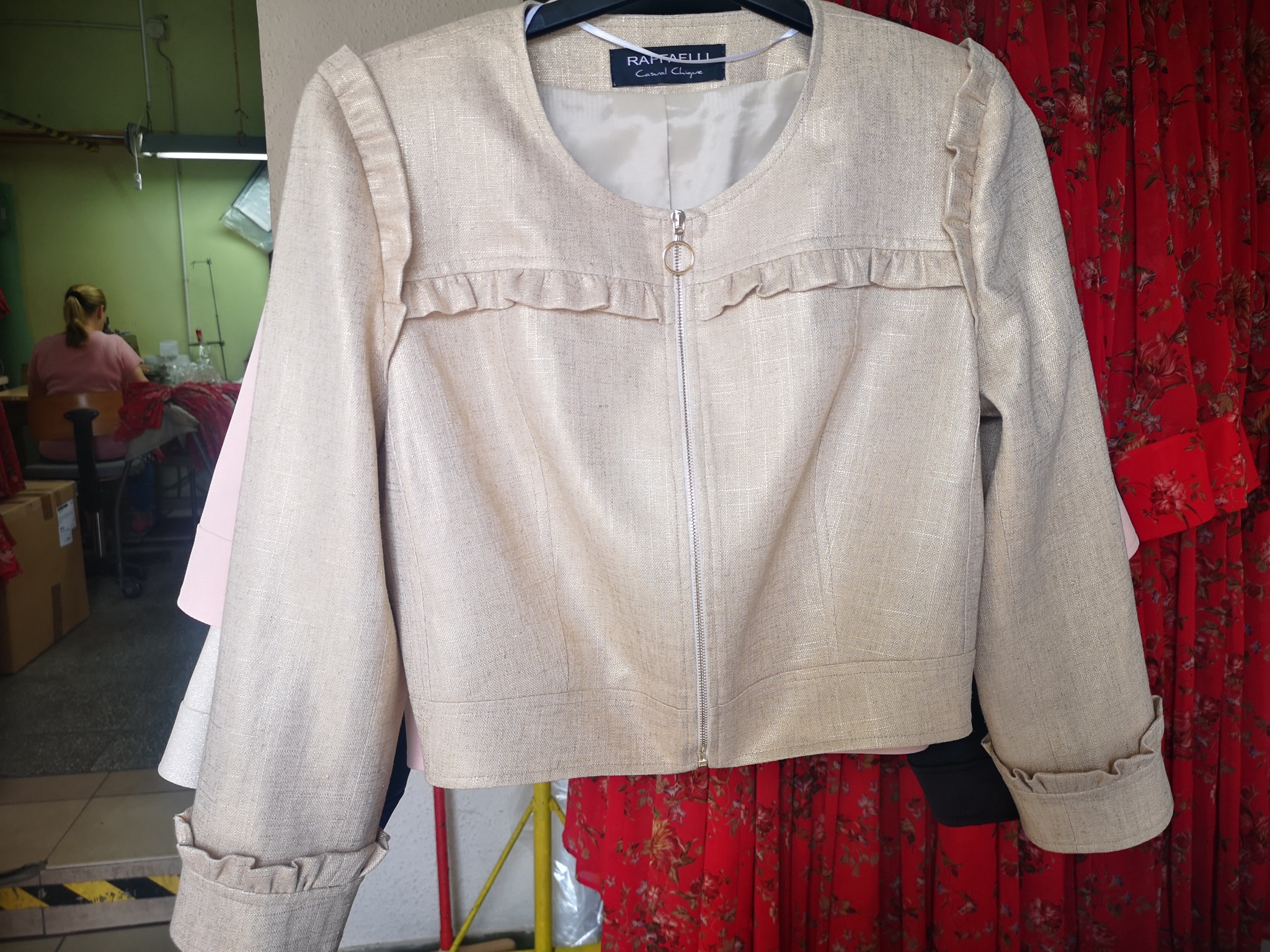 Beżowa damska kurtka z lnu z falbankami, zapinana na zamek błyskawiczny, wisząca na wieszaku w szwalni, widoczne inne ubrania i stanowisko pracy krawcowej w tle.