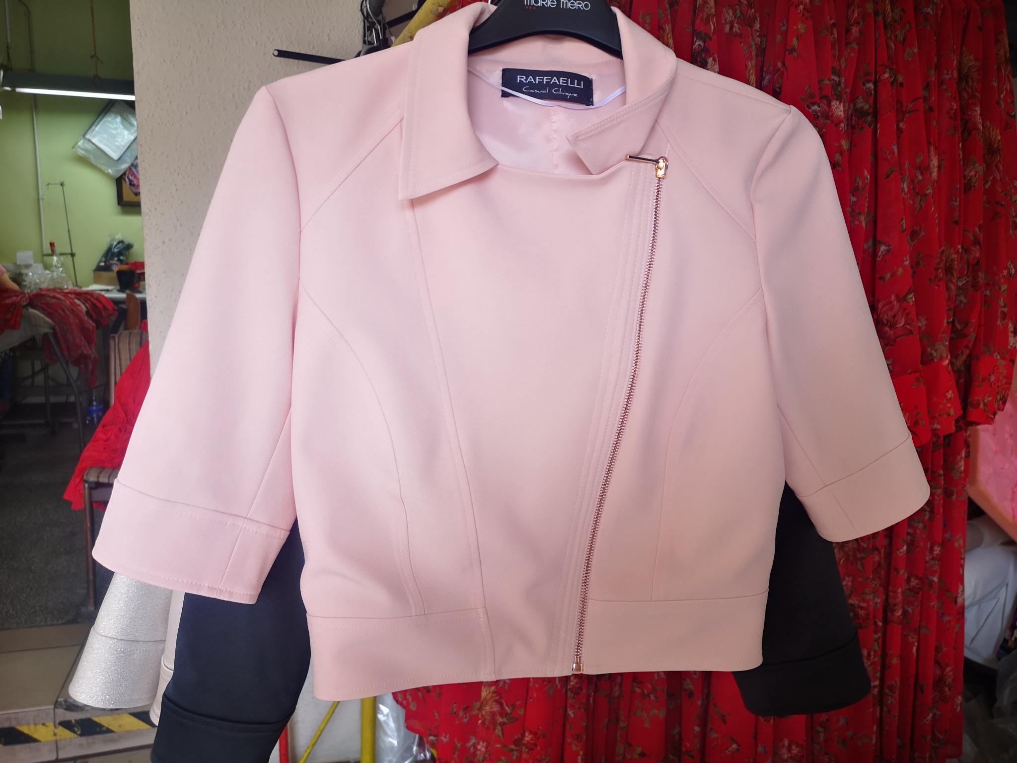 Krótki, pudrowo-różowy żakiet z asymetrycznym zamkiem błyskawicznym w kolorze różowego złota, wiszący na wieszaku na tle wzorzystej czerwonej sukienki.