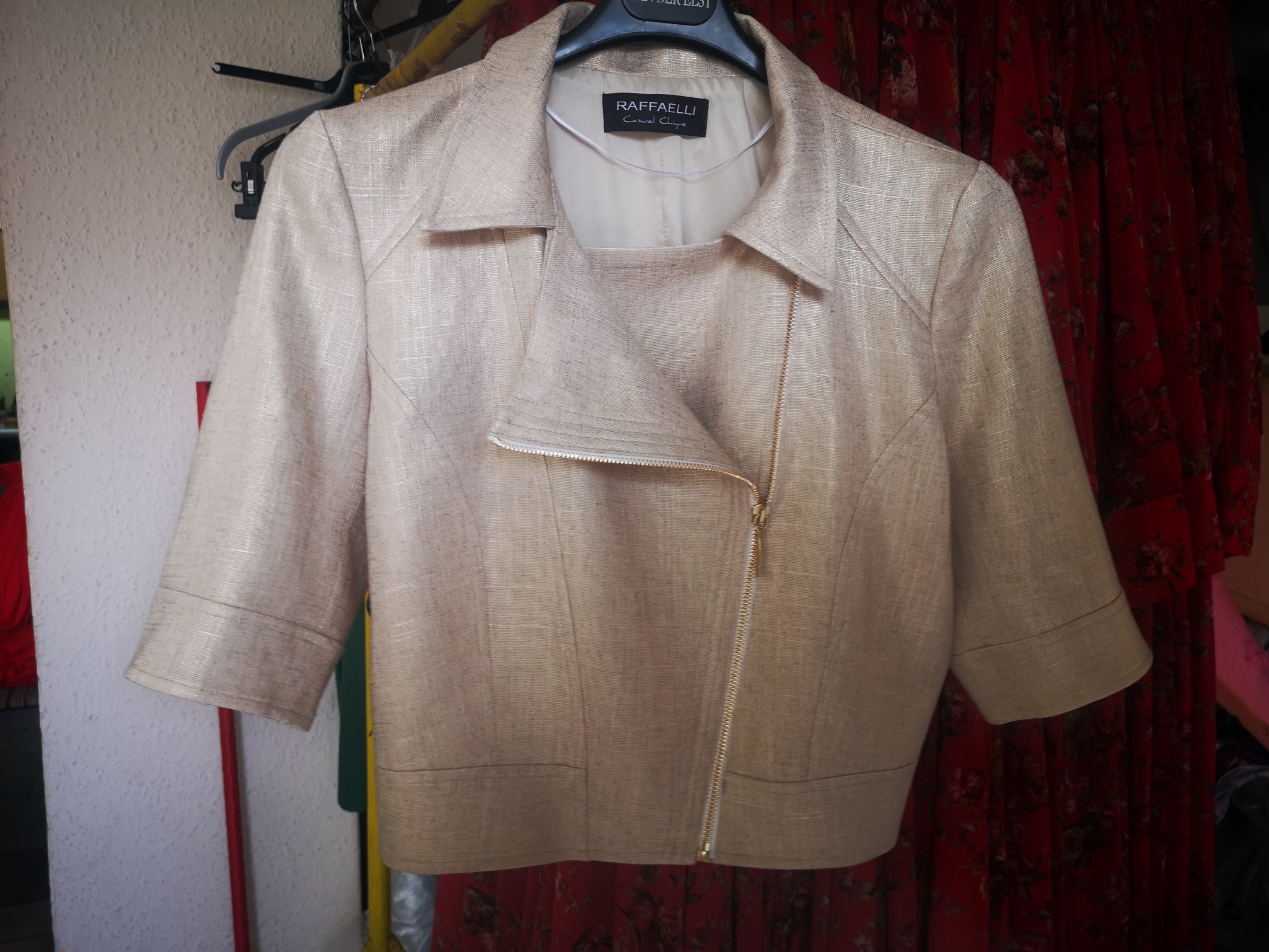 Krótki, beżowy żakiet z asymetrycznym zapięciem na zamek, wiszący na wieszaku na tle wzorzystej, czerwonej zasłony.