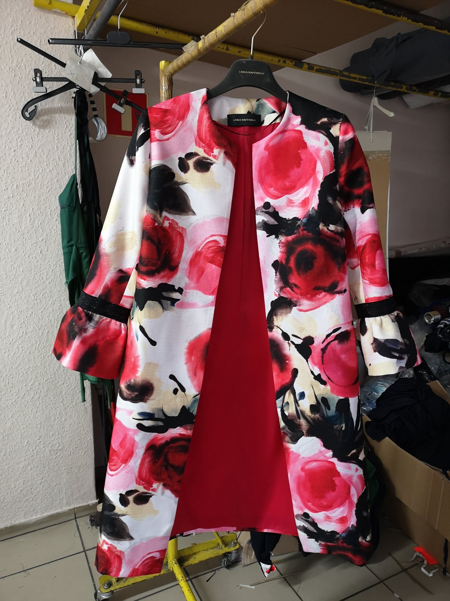 Płaszcz damski z kwiecistym wzorem w odcieniach czerwieni, bieli i czerni, wiszący na wieszaku, z widoczną czerwoną podszewką i falbanami na rękawach, w tle widoczne inne ubrania i elementy...