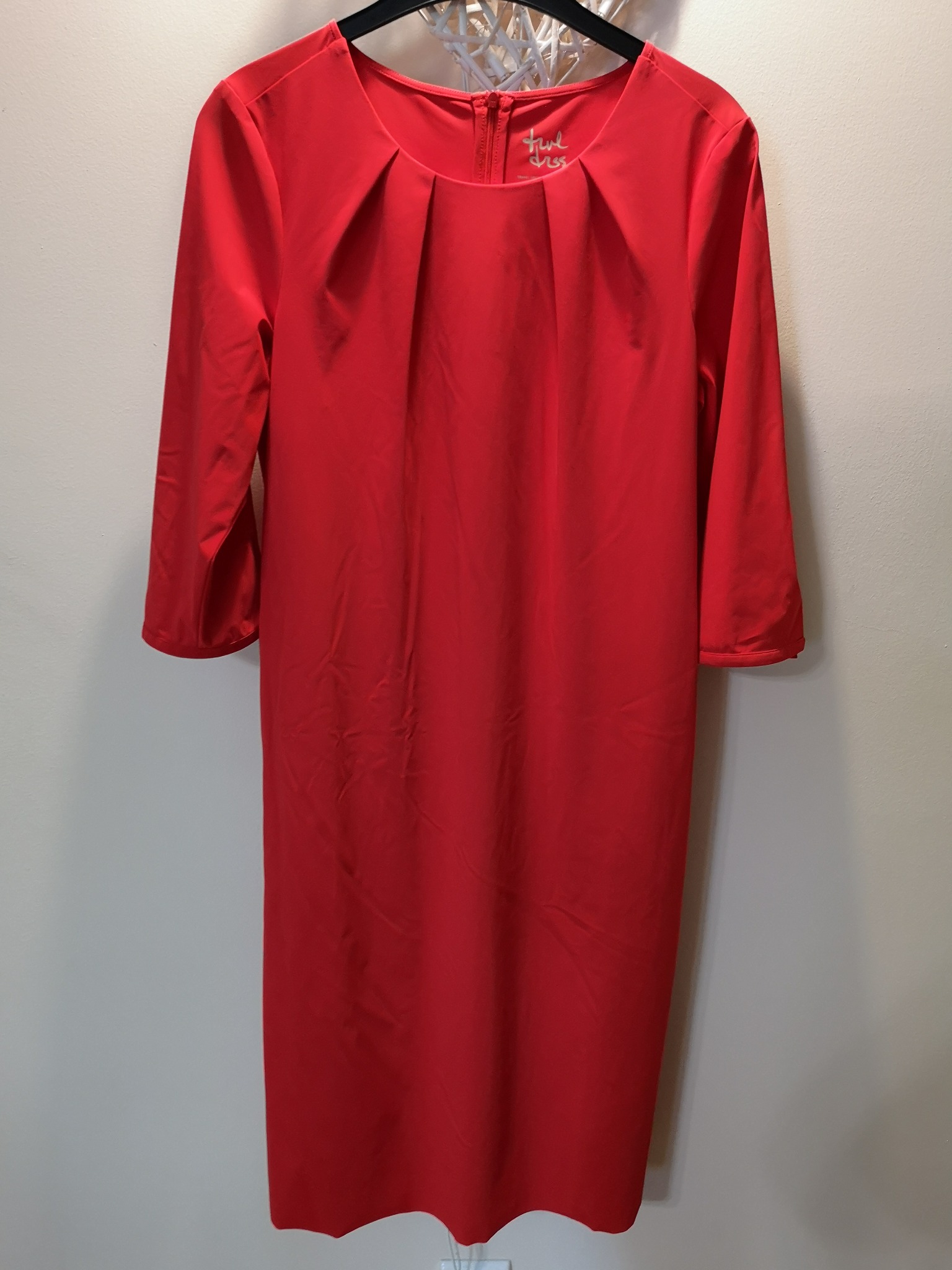 Czerwona sukienka z trzema czwartymi rękawami, wisząca na czarnym wieszaku przed kremową ścianą, widoczny zamek błyskawiczny i metka z napisem.