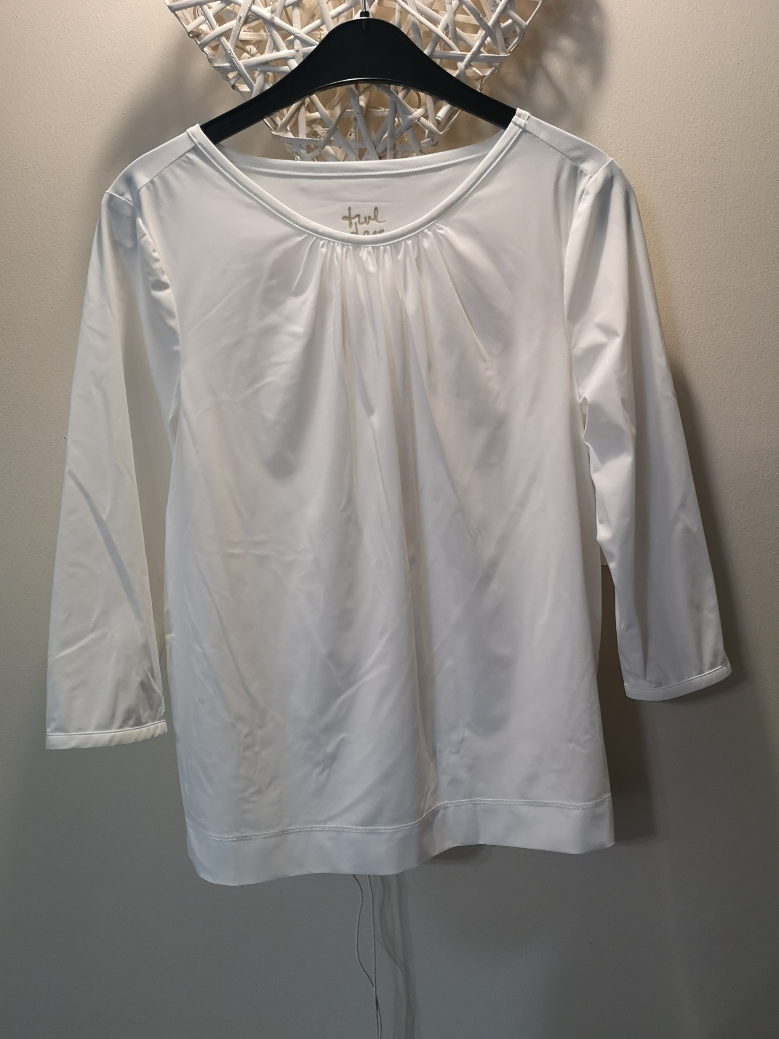 Prosta, biała bluzka z rękawami 3/4, wisząca na czarnym wieszaku na tle kremowej ściany z dekoracją w kształcie gwiazdy.