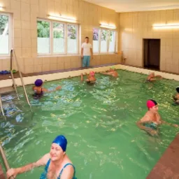 Kryty basen z zieloną wodą, gdzie grupa osób w czepkach pływa i ćwiczy, a mężczyzna stoi przy oknie w tle.