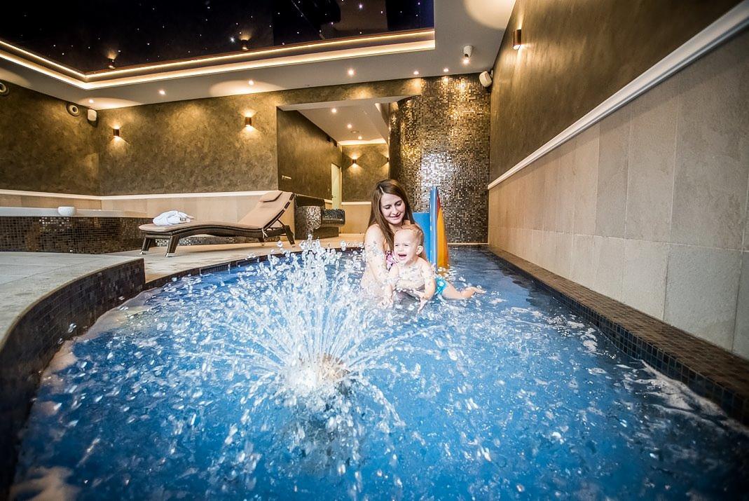 Wnętrze luksusowego spa z basenem, w którym kobieta bawi się z małym dzieckiem, fontanna tryska wodą, w tle leżanka i ściana z mozaiką.