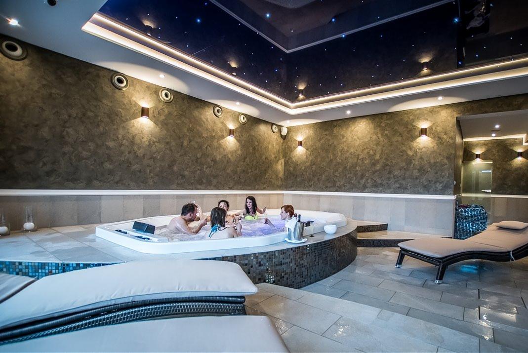 Grupa osób relaksuje się w luksusowym jacuzzi w pomieszczeniu z dekoracyjnym oświetleniem sufitowym imitującym gwiazdy i ścianami z efektem kamienia. Leżanki obok wanny.