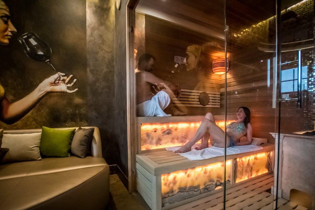 Nowoczesna strefa relaksu z przeszkloną kabiną, podświetlane drewniane ławy, mural z kobietą i kieliszkiem, oraz skórzana sofa z poduszkami.