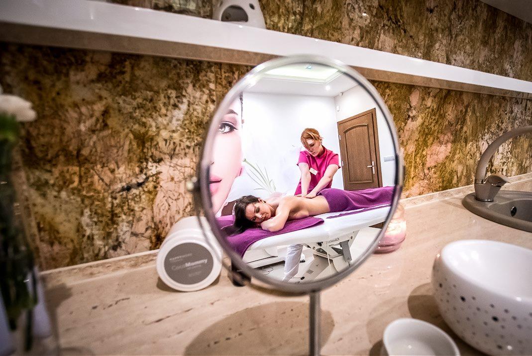 Masaż pleców w salonie spa, widoczny w odbiciu okrągłego lustra na tle marmurowej ściany i kosmetyków.