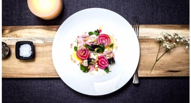 Elegancko podana sałatka z buraków z serem kozim, orzechami piniowymi i świeżymi ziołami na białym talerzu, ustawionym na drewnianej desce z dodatkiem świeczki, soli, pieprzu i kwiatów.