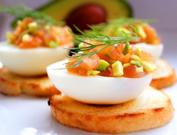 Eleganckie przystawki: Połówki jajek na grzankach, wypełnione kostkami łososia i awokado, udekorowane świeżym koperkiem, na białym talerzu z rozmytym awokado w tle.